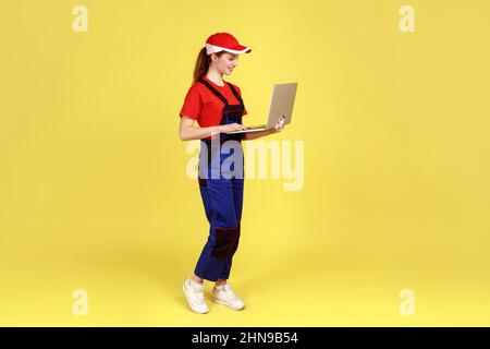 Portrait de la femme souriante travaillant sur ordinateur portable, traite les commandes en ligne, exprime des émotions positives, porte une combinaison et une casquette rouge. Studio d'intérieur isolé sur fond jaune. Banque D'Images