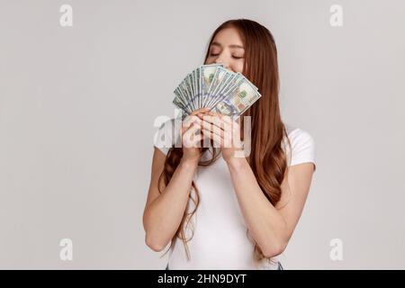 Portrait d'une femme avaricieuse à cheveux foncés qui sent des billets de banque en dollars, qui profite des gains de loterie, des profits importants et de la vie riche, portant un T-shirt blanc. Prise de vue en studio isolée sur fond gris. Banque D'Images