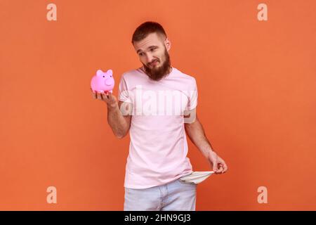 Portrait d'un pauvre barbu tenant la porcgybank à la main et montrant ses poches vides, frustré et contrarié, portant un T-shirt rose. Studio d'intérieur isolé sur fond orange. Banque D'Images