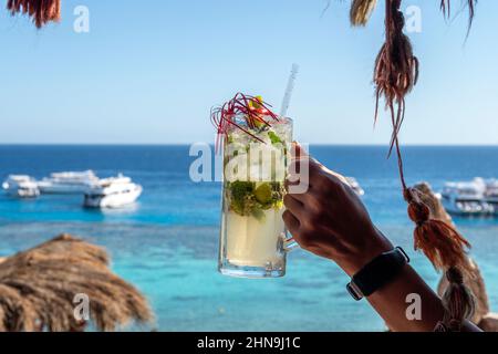 Main de femme tenant un verre de mojito frais avec de la glace, de la menthe, du citron vert et des citrons. Toile de fond de la plage. Couleurs bleu et vert améliorées. Égypte Banque D'Images