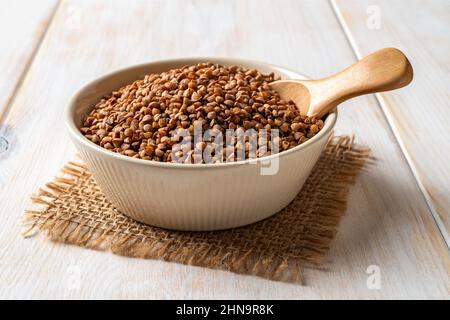 Bol en céramique beige rempli de gruaux de sarrasin cru et d'une cuillère sur une table en bois blanc. Cuisson du porridge de sarrasin. Concept de régime sans gluten. Banque D'Images