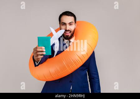 Homme d'affaires souriant et heureux portant un anneau en caoutchouc orange, un avion en papier et un passeport, planifiant la visite, portant un costume de style officiel. Prise de vue en studio isolée sur fond gris. Banque D'Images