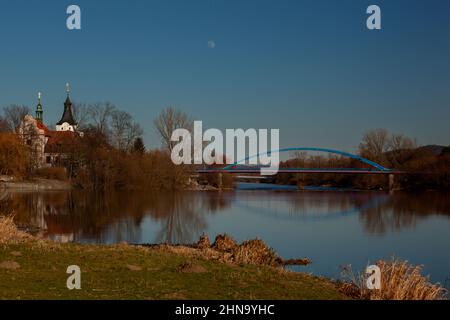 La rivière est calme et reflète le pont et l'église. L'herbe verte est au premier plan. Le ciel est bleu sans nuages. La lune est presque pleine. Banque D'Images