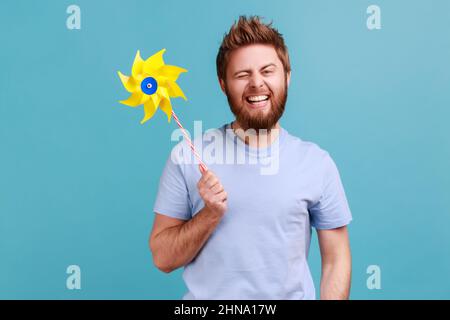 Portrait de positif ravie beau barbu homme souriant sans souci et se want à l'appareil photo, tenant le moulin à vent de papier, le jouet de roue de PIN sur bâton. Studio d'intérieur isolé sur fond bleu. Banque D'Images