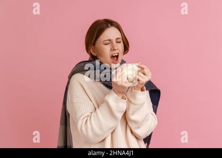 Portrait d'une femme blonde malsaine et malsaine éternuant, attrapant le froid, se tenant enveloppé dans un foulard, souffrant de grippe, portant un chandail blanc. Studio d'intérieur isolé sur fond rose. Banque D'Images