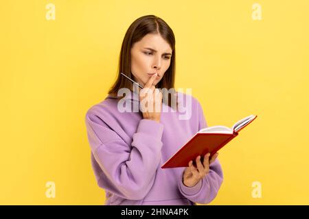 Femme attentionnée debout avec un carnet en papier et un stylo, regardant loin avec l'expression pensive, faisant la liste des tâches, portant le pull à capuche violet. Studio d'intérieur isolé sur fond jaune. Banque D'Images