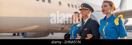 Un pilote homme excité marchant avec deux agents de bord en uniforme bleu devant un avion dans le terminal au coucher du soleil Banque D'Images