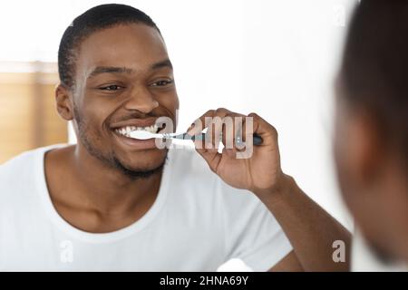 Concept de routine du matin. Happy Black Man se brossant les dents près du miroir dans la salle de bains, Banque D'Images