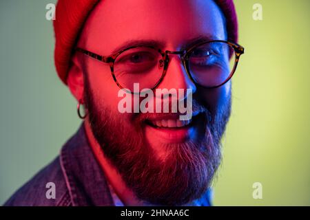 Gros plan portrait de l'homme hippster heureux à barbe regardant l'appareil photo avec un sourire crasseux, optimisme, succès, portant un bonnet beanie rouge. Studio d'intérieur isolé sur fond de néon coloré. Banque D'Images