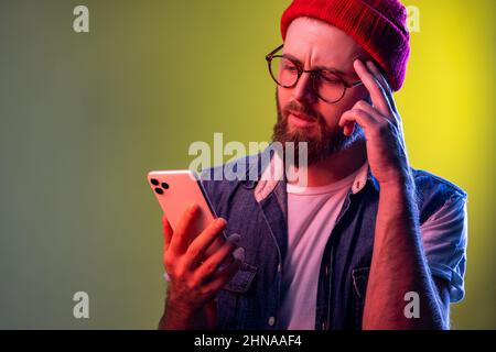 Homme hippster à la main, pensant à la mise à jour de logiciels, pensant à choisir des tarifs adaptés. Studio d'intérieur isolé sur fond de néon coloré. Banque D'Images