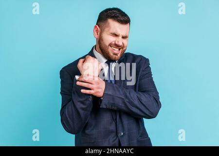 Portrait d'un homme d'affaires malade portant un costume officiel debout avec de la grimace de douleur, massant mal au poignet, souffrant de blessure à la main ou d'entorse. Studio d'intérieur isolé sur fond bleu. Banque D'Images
