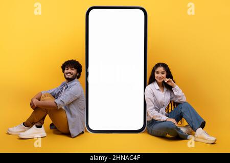 Un couple indien enthousiaste assis près d'un smartphone géant avec écran blanc vierge, démontrant l'espace de copie de votre annonce Banque D'Images