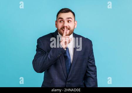 Portrait d'homme d'affaires portant un costume officiel tenant le doigt près des lèvres montrant le geste shh, gardant les secrets se préparer à la surprise. Studio d'intérieur isolé sur fond bleu. Banque D'Images