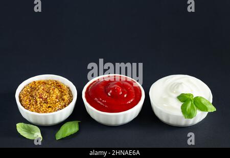 Ensemble de sauces populaires : ketchup, moutarde de dijon et mayonnaise dans de petits bols blancs sur fond noir. Copier l'espace Banque D'Images