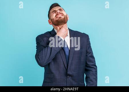 Douleur à la gorge. Portrait d'un homme d'affaires malade malheureux portant un costume officiel touchant son cou, souffrant de maux de gorge, d'infection virale ou de symptômes de grippe. Studio d'intérieur isolé sur fond bleu. Banque D'Images