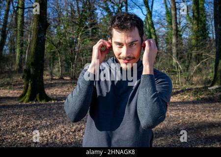 Le coureur écoute de la musique tout en s'étirant dans les bois. Portrait d'un beau homme aux yeux bleus et d'une moustache qui s'entraîne dans le parc. Banque D'Images