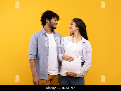 Heureux couple indien s'attendant à bébé, posant sur fond jaune, embrassant et regardant l'un l'autre Banque D'Images