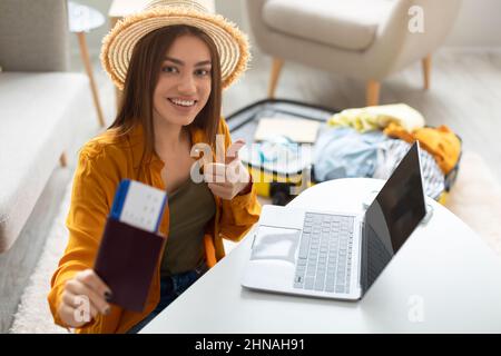 Une jeune femme heureuse avec un passeport et des billets à l'aide d'un ordinateur portable, montrant le pouce vers le haut, recommandant l'agence de voyage Internet à la maison Banque D'Images
