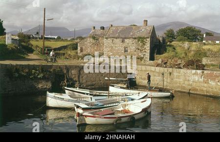 1960s, photo historique de cette époque de petits bateaux à rames amarrés dans le port d'Annalong, Co Down, Irlande du Nord, Royaume-Uni, avec deux lads debout en un et un homme local assis avec son vélo sur la terre sèche au-dessus. Village balnéaire pittoresque et paisible, Annalong se trouve au pied des montagnes de la Mourne. Banque D'Images