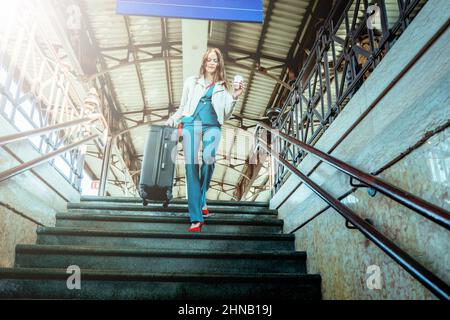 Une jeune femme descend les escaliers de la gare avec un sac à roulettes - une femme d'affaires descend les marches du terminal avec ses bagages Banque D'Images