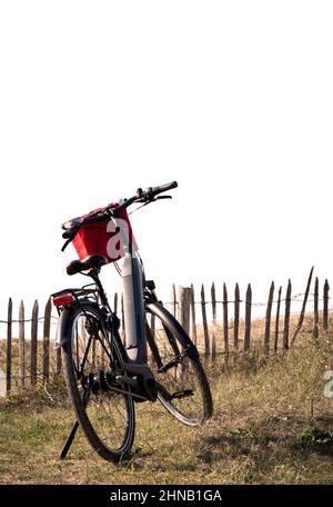 Vélo électrique noir avec un panier rouge garé dans l'herbe contre la clôture sur fond blanc pour le texte de copie Banque D'Images