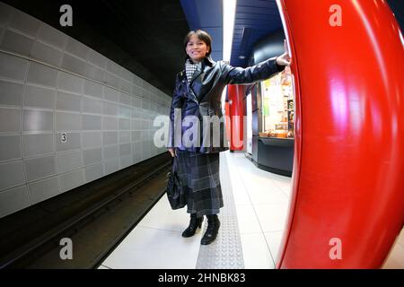 Une femme dans une veste en cuir et une jupe à carreaux se tient sur une plate-forme de métro moderne, appuyée contre une colonne structurelle rouge vif. Banque D'Images