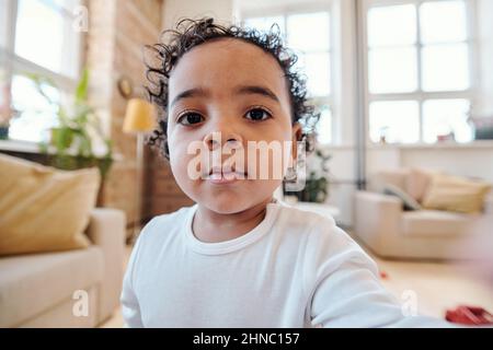 Selfie de bébé garçon de race mixte mignon avec des cheveux mauves foncés portant une chemise en jute assis dans le salon Banque D'Images