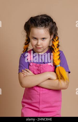 Offensé petite fille ayant des tresses jaunes kanekalon regardant l'appareil photo de l'Alaska avec les bras pliés portant la combinaison rose et le t-shirt violet sur beige Banque D'Images