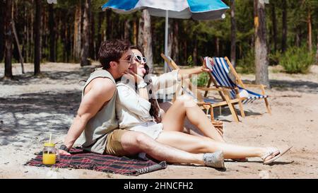 Couple positif en lunettes de soleil prenant le selfie sur la couverture sur la plage Banque D'Images