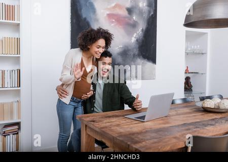 une femme afro-américaine heureuse enceinte agite la main sur un ordinateur portable près de son mari, montrant le pouce vers le haut Banque D'Images