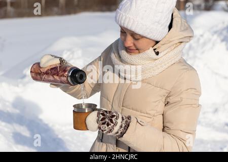 Une belle jeune femme dans une veste en duvet et des mitaines verse une boisson chaude dans une tasse d'un thermos dans la nature lors d'une belle journée d'hiver. Mise au point sélective. Por Banque D'Images