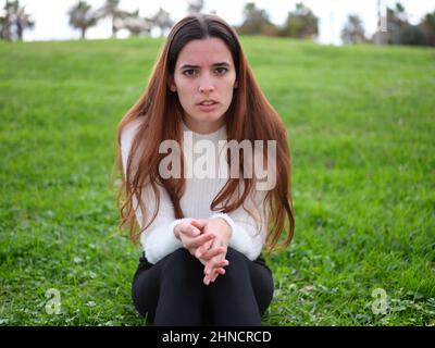 Une jeune femme en colère dans le parc assise sur l'herbe avec ses mains ensemble regardant la caméra. Banque D'Images