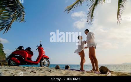 Trajet en scooter. Un couple charmant sur une moto rouge vêque de vêtements blancs sur une plage de sable. Mariage juste marié personnes marchant près de palmiers tropicaux, mer. Lune de miel au bord de l'océan. Asie Thaïlande. Location de motos. Banque D'Images