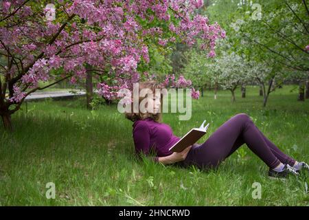 jeune femme, étudiante, lit le livre avec intérêt, couché sous l'arbre le jour du printemps dans le parc. filles - adolescent 17 ans avec livre sur l'herbe. préparation Banque D'Images