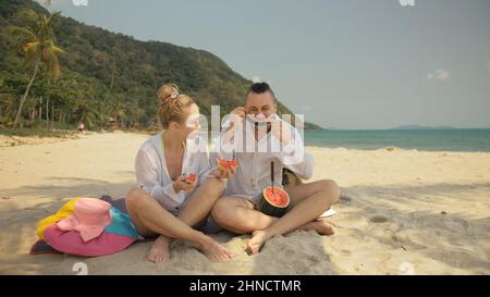 Le couple d'amour gai tenant et mangeant des tranches de pastèque sur le sable tropical plage mer. Amoureux romantiques deux personnes caucasiens passent le week-end d'été en plein air. Chapeau, sac à dos chemise blanche vêtements de plage. Banque D'Images