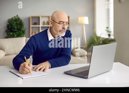 Homme senior assis à un bureau avec un ordinateur portable, participant au séminaire en ligne et prenant des notes d'étude Banque D'Images