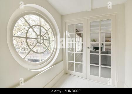 Mur blanc avec fenêtre ronde dans le couloir lumineux de l'appartement moderne avec portes vitrées fermées du salon avec canapé Banque D'Images