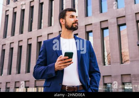 Ci-dessous le contenu barbu homme entrepreneur en tenue habillée regarder loin tandis que la messagerie texte sur smartphone près de bâtiment moderne de plusieurs étages dans la rue Banque D'Images