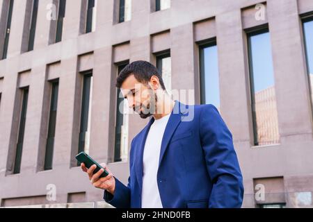 Ci-dessous le contenu barbu homme entrepreneur en tenue habillée regarder loin tandis que la messagerie texte sur smartphone près de bâtiment moderne de plusieurs étages dans la rue Banque D'Images