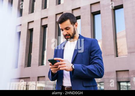Ci-dessous le contenu barbu homme entrepreneur en tenue habillée regarder loin tandis que la messagerie texte sur smartphone près de bâtiment moderne de plusieurs étages dans la rue Banque D'Images