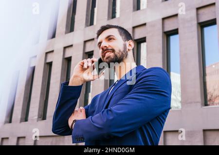 Ci-dessous le contenu barbu homme entrepreneur en tenue formelle regardant vers le haut tout en ayant la conversation téléphonique près de bâtiment moderne de plusieurs étages sur la rue de ci Banque D'Images