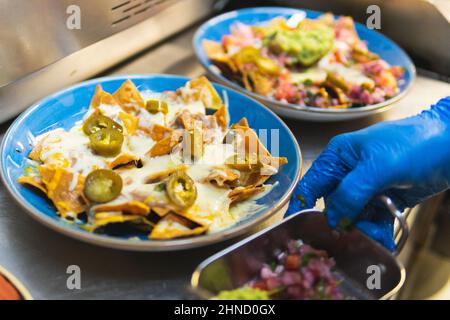 Croise le chef méconnaissable dans un gant en latex avec un récipient de remplissage près des assiettes avec des nachos recouverts de tranches de jalapeno chaudes Banque D'Images