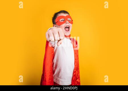 Enfant dans des vêtements rouges superhéros et masque tirant le bras tout en se tenant sur fond jaune dans le studio et l'avion d'imagerie Banque D'Images