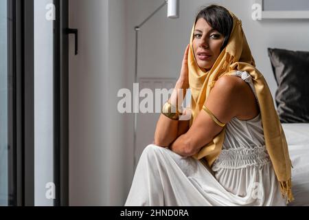 Jolie femme musulmane en tenue blanche et hijab jaune traditionnel regardant l'appareil photo tout en étant assise sur le lit dans la chambre lumineuse Banque D'Images