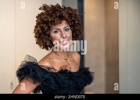 Belle femme ethnique dans un élégant vêtement noir avec des cheveux courts et bouclés souriant et regardant l'appareil photo à la maison Banque D'Images