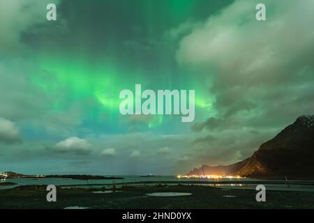 Paysage pittoresque de petite ville côtière avec des maisons illuminées sur la mer près des montagnes rocheuses contre ciel nuageux avec lumières polaires lumineuses dans Norw Banque D'Images