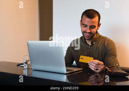 Homme barbu positif regardant la carte de crédit en plastique tout en effectuant le paiement en ligne sur netbook moderne à la table dans la pièce légère Banque D'Images
