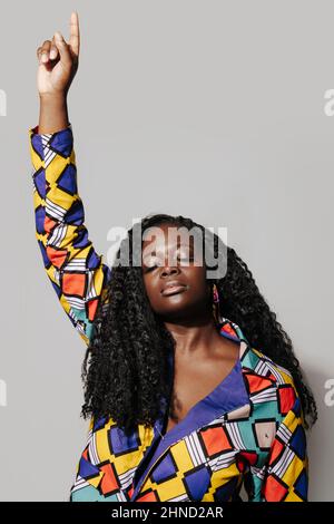 Femme afro-américaine pensive avec cheveux bouclés dans une tenue africaine élégante debout sur fond blanc. Elle est avec les yeux fermés pointant le doigt vers le haut Banque D'Images