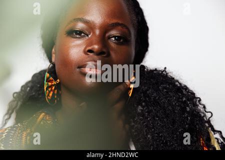 Portrait d'une jeune femme aux courbes portant des vêtements africains élégants, regardant l'appareil photo tout en étant debout dans une pièce lumineuse. Elle a des plantes non concentrées dans le foregrou Banque D'Images