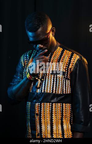 Jeune homme dans une élégante tenue africaine et des lunettes de soleil regardant loin tout en se tenant sur fond noir dans le studio avec des lumières en gel colorées Banque D'Images